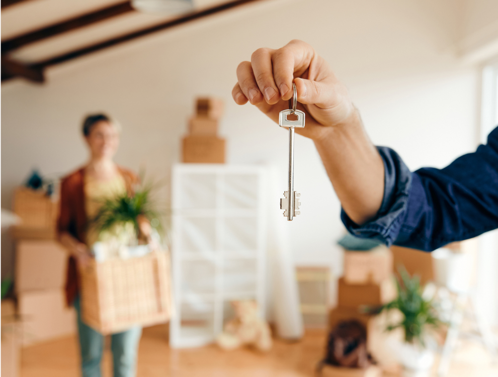 A person holding a key, with a woman in the background smiling and moving items, symbolizing a successful home sale through Zown.