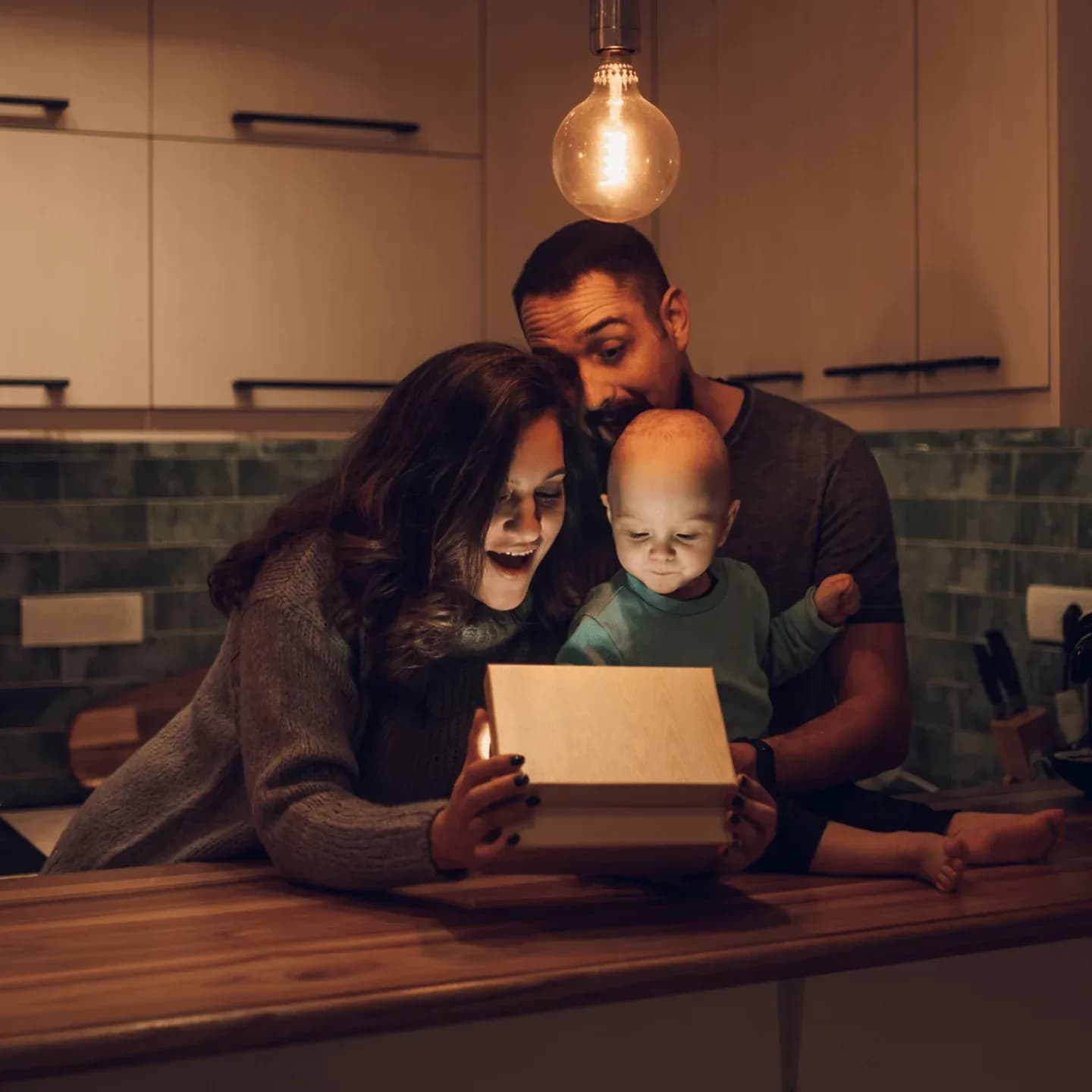 Warmly lit kitchen scene of a young family gathered around a glowing wooden box on a countertop — a smiling mother and father leaning in close as their baby looks curiously at the light inside, creating a cozy, hopeful moment that evokes surprise, possibility, and new beginnings.