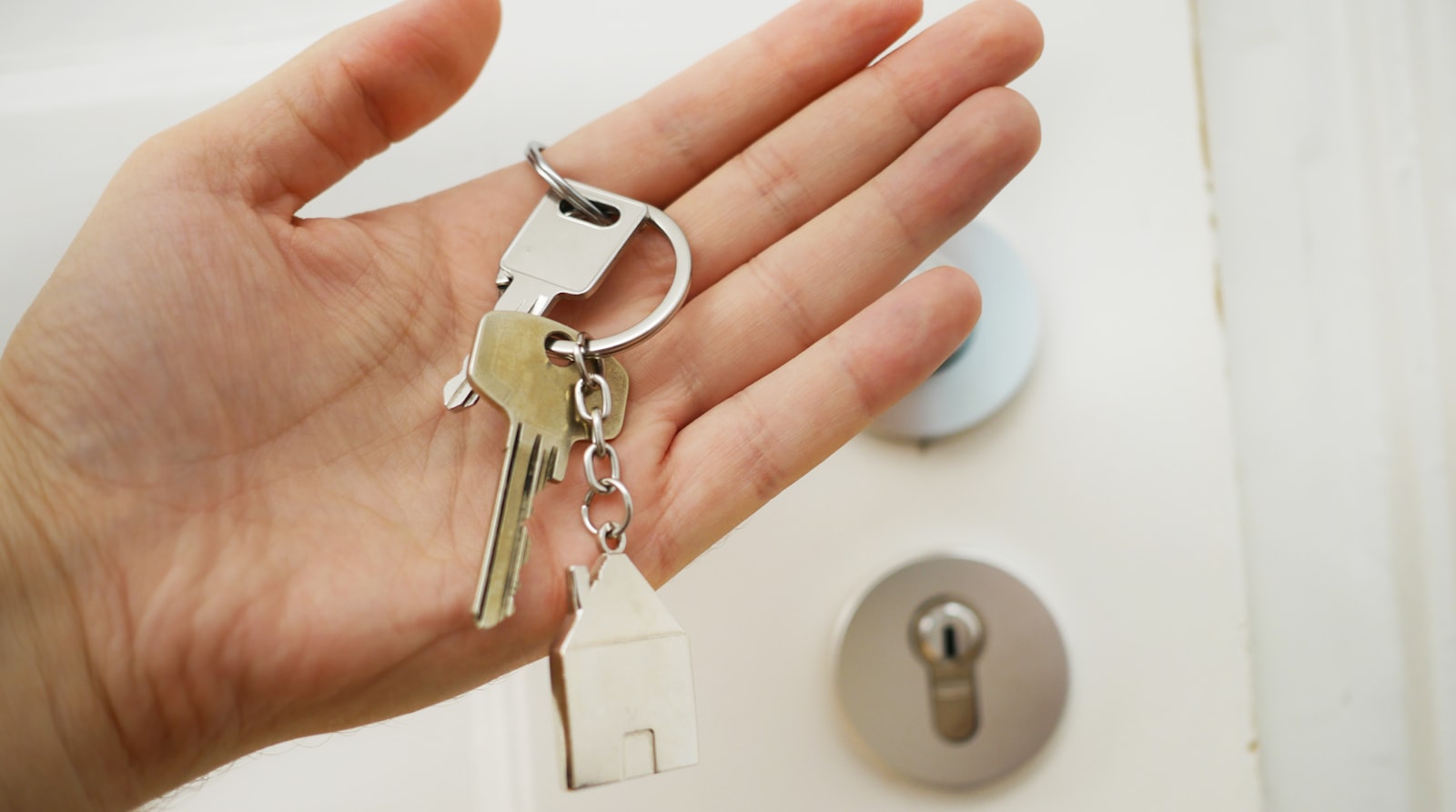 An open hand holding a set of house keys with a small house keyring in front of a white door